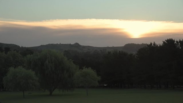 Sundown on a landscape with mountains on the back, a lot of trees, green grass on a cloudy day with sunrays on the image