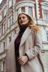 Attractive young blonde woman in casual coat looking at the camera on the city street. Beautiful architecture building on background. Portrait shot