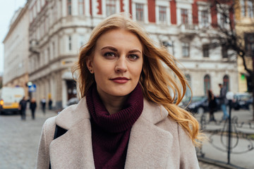 Close up portrait of young beautiful woman with blonde hair looking at the camera while standing on the city street on spring day.