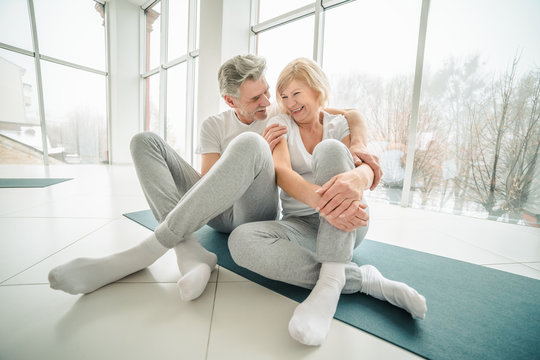 Sporty senior couple sitting on the mat in the white fitness gym looking each other and talking. Sports and healthy lifestyle. - Powered by Adobe