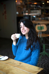 Portrait of a beautiful smiling young lady drinking coffee and looking at camera while sitting at table in a cafe