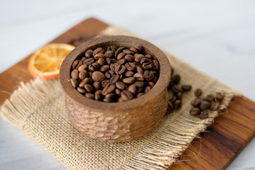 Closeup of coffee beans in wooden rustic bowl on bright background decorated with orange slice.