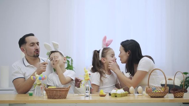 Happy and positive family: mother, father and two kids are smiling on the white background. They ar playing with paint-brushes. Mommy gives a little kiss to her lovely daughter. Easter holiday concept
