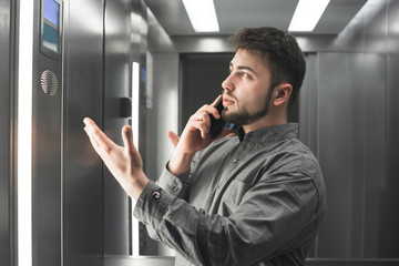 Serious businesswoman explaining something by the phone in the elevator wearing grey shirt. Bearded man employee is peacefully talking on her smartphone and actively gestures