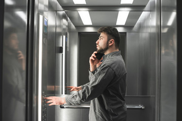Young european man office worker is looking at the number of the floor in the lift and talking on the phone. Businessman is over the phone in the elevator pressing the button.