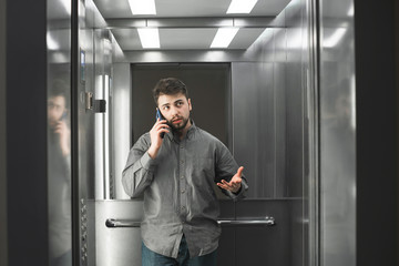 Serious businesswoman explaining something by the phone in the elevator wearing grey shirt. Bearded man employee is peacefully talking on her smartphone and actively gestures