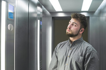 Portrait of a bearded adult man wearing a gray shirt stands in a lift with a serious face and looks side by side on the number on the floor. Serious man in the elevator.