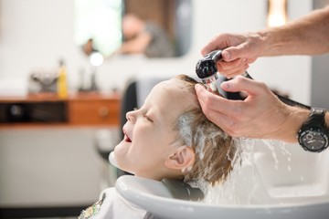 Hairdresser washing your child's hair with shampoo in barbershop. Joyful child gets pleasure from...