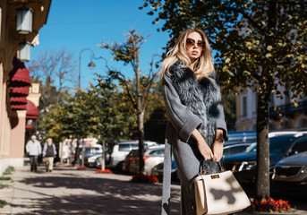Obraz na płótnie Canvas Beautiful young woman in sun glasses and fur coat posing with luxury bags and accesories while standing walking outdoors. Female fashion city lifestyle outfit spring or autumn shopping concept