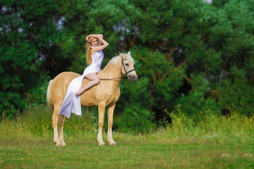 Young rider woman blonde with long hair in a white dress with a train posing and jumping on a palamino horse against a field and forest background
