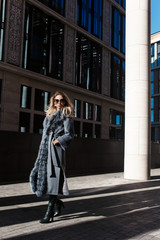 Obraz premium Stylish woman in rich fur coat and sunglasses in modern city street columns background, autumn fashion trend, green boots, urban style, accessories, legs, footwear. Autumn or spring fashion shopping