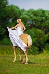 Young rider woman blonde with long hair in a white dress with a train posing and jumping on a palamino horse against a field and forest background