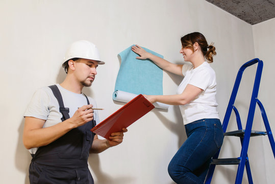 Funny couple, woman worker and her boss in helmet wallpapering the walls using a metal ladder. Home renovation and moving to new house concept