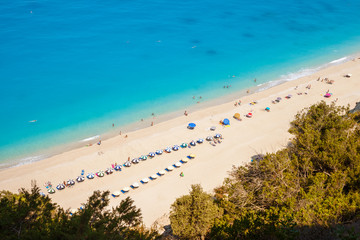 Aerial view of Egremni beach, Lefkada, Greece, on a sunny Summer day, with turquoise waters and beach umbrellas along the shore line. Unrecognizable people sun bathing and swimming.