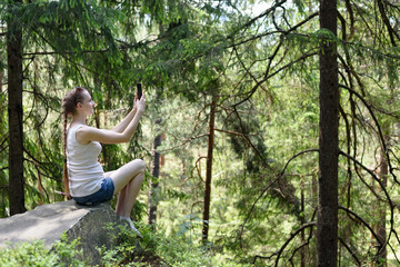 Dreaming beautiful girl sitting on a stone and making selfie on a smartphone surrounded by coniferous forest on a sunny day