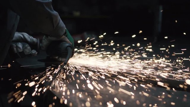 Metal industry worker in a dark workshop using flex saw to grind metal artwork on working table.
