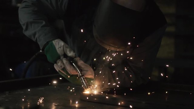Metal industry worker welding iron item on steel table in dark workshop with full equipment on.