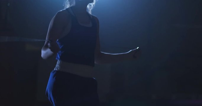 Young beautiful woman professional boxer in training in the hall jumps on a rope in the light of the contra in a dark room in blue clothes