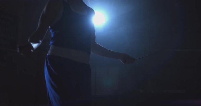 Cardio training for fighters. Young beautiful woman professional boxer in training in the hall jumps on a rope in the light of the contra in a dark room in blue clothes