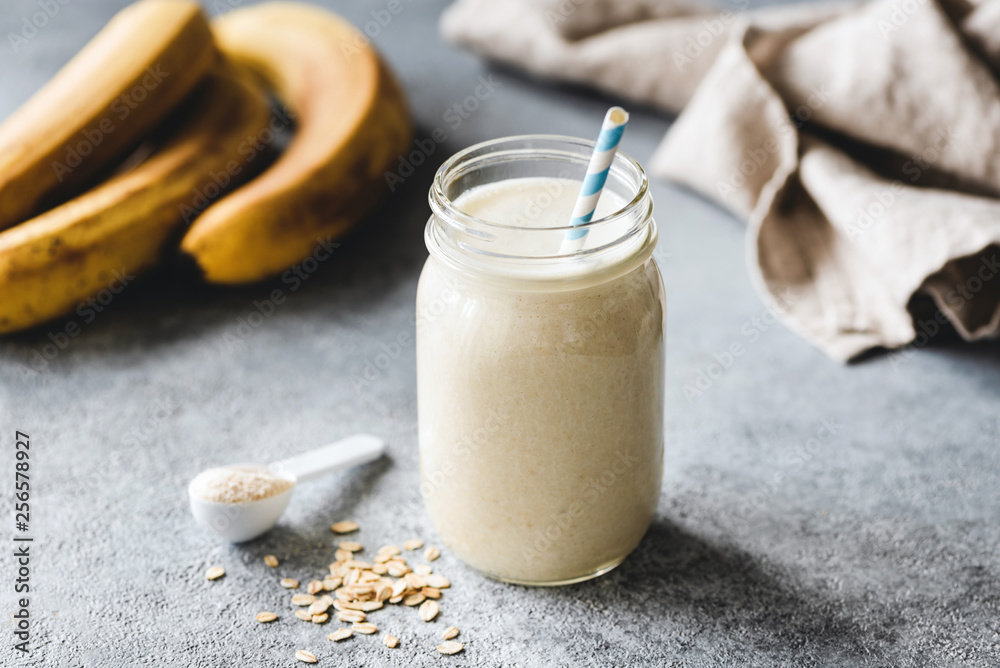 Wall mural Protein banana smoothie in jar, protein scoop, oats and linen textile on table. Healthy vegan or vegetarian drink for sporty people