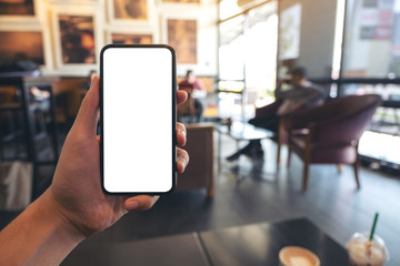 Mockup image of a hand holding and showing black mobile phone with blank white screen in cafe