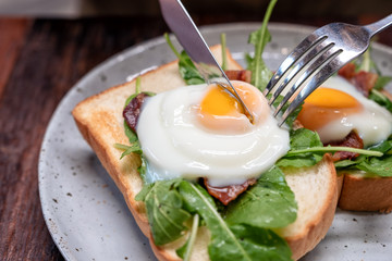 A woman eating breakfast sandwich with eggs, bacon and sour cream by knife and fork in a plate on wooden table