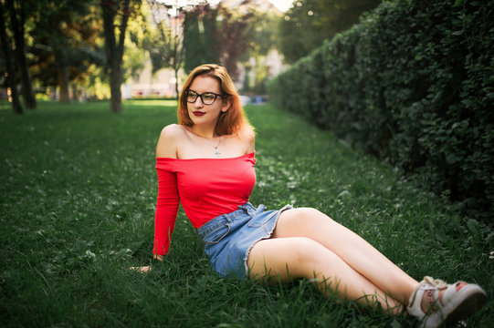 Attractive redhaired woman in eyeglasses, wear on red blouse and jeans skirt posing at green park.