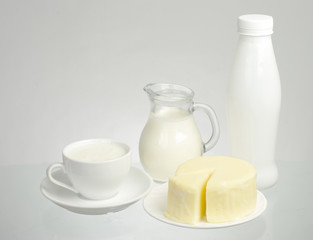 A cup of milk, a jug of milk, a bottle of milk and a piece of cheese on a white background.