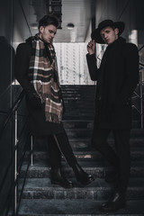 Young stylish men posing on steps near city center. Wearing in black coat, scarf, hat, pants, boots. Street style men model tests.