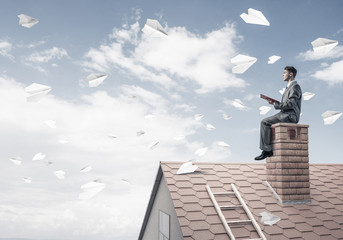 Man on brick roof reading book and paper planes flying in air