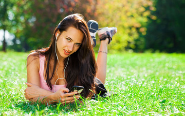 Young woman in dress lies on green grass and listens to music in headphones through smartphone.