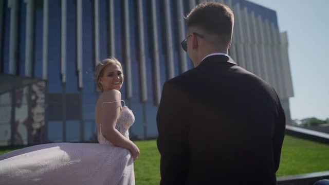 beautiful couple of newlyweds walking in a park with a beautiful white building. Young groom in a black suit and bride with long hair in a wedding dress holding hands. A beautiful couple of newlyweds 