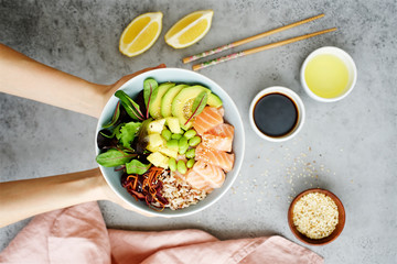 Girls' hands holding salmon poke bowl Salmon poke bowl with salad, avocado, edamame beans, pineapple, sesame and brown rice Traditional Hawaiian dish