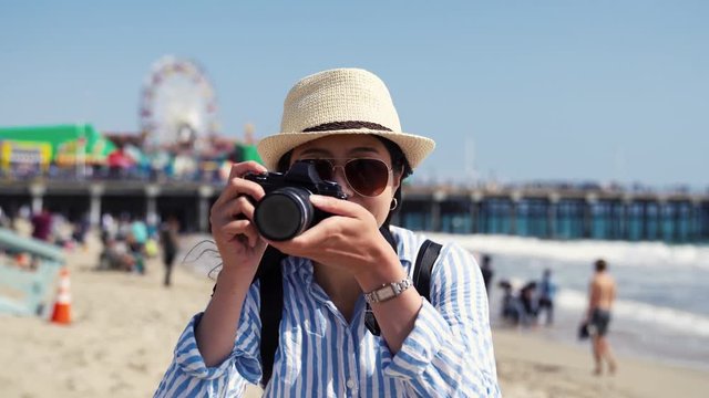 happy asian female photographer holding slr camera taking photo picture of nature sandy beach bay summer view Santa Monica Pier. people relaxing having fun in background with water sea and waves.