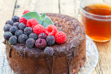 Homemade chocolate cake decorated with black and red raspberries on glass plate with cup of tea.
