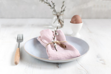 Fototapeta na wymiar Easter table setting idea, minimal decoration - pink napkin, fork, coloured egg in white egg cup, willow banch, white wooden table