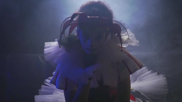 Girl clown in a dress, a hat and makeup is sitting in the dark room under the spotlight