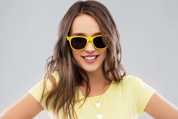 Naklejka premium summer, accessory and people concept - smiling young woman or teenage girl in yellow t-shirt and sunglasses over grey background