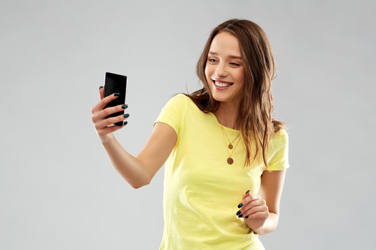 technology and people concept - smiling young woman or teenage girl in blank yellow t-shirt taking selfie by smartphone over grey background