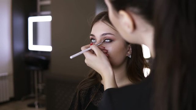 Visagist correcting the low eyelid with a brush and black pencil. Checking her work in mirror reflection