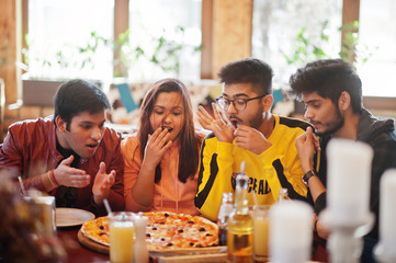 Group of asian friends eating pizza during party at pizzeria. Happy indian people having fun together, eating italian food and sitting on couch. Shocked and surprise faces.