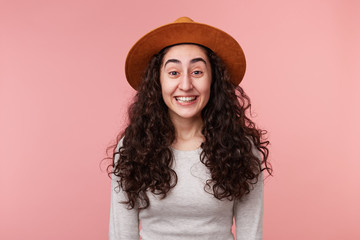 Portrait of young famale with dark curly hair, wears casual white longsleeve, with a hat on head, smiles broadly, isolated over pink background. Positive emotion concept.