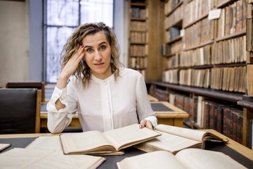 Girl in the reading room of the library to prepare for the exam holding his head with a bunch of books on the table