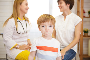 Little boy with teddy bear is looking at the camera. Female doctor and mother on background
