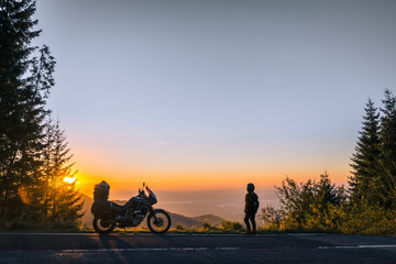 girl in full motorcycle equipment, stands on the side of the road over a cliff and looks into the distance at beautiful sunset in the mountains. Adventure motorcycle, Transfagarasan, Romania