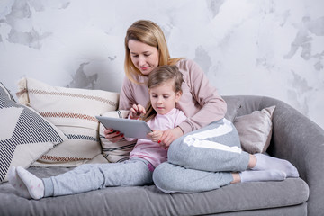 Obraz na płótnie Canvas Beautiful young mother and her little daughter are watching movies together and playing on the tablet while sitting on the sofa. Mom hugs daughter. Maternal care and love. Horizontal photo