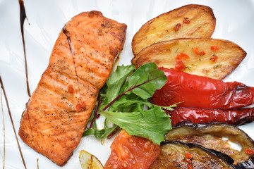 A piece of fried fish and grilled vegetables in a white plate in the restaurant. Fish and baked vegetables eggplant, pepper and tomatoes. Closeup