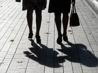 Silhouettes of two women walking down the street. Girls in skirts and shoes on high heels outdoors, shadows on pavement, concept for female friendship, fashion, dramatic life stories