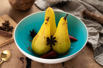 Caramelized poached pears - with anis, red chili pepper, cinnamon spices in syrup on the blue plate on a wooden table. Delicious cooked pear dessert for holiday. Stewed pears in syrup with anise dust