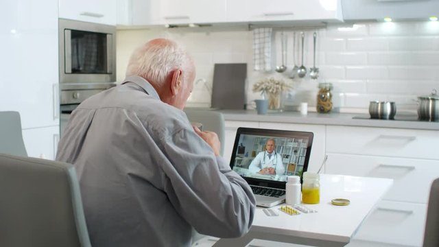 Medium shot of sick elderly Caucasian man looking at computer screen and listening to doctor recommendations concerning his health state
